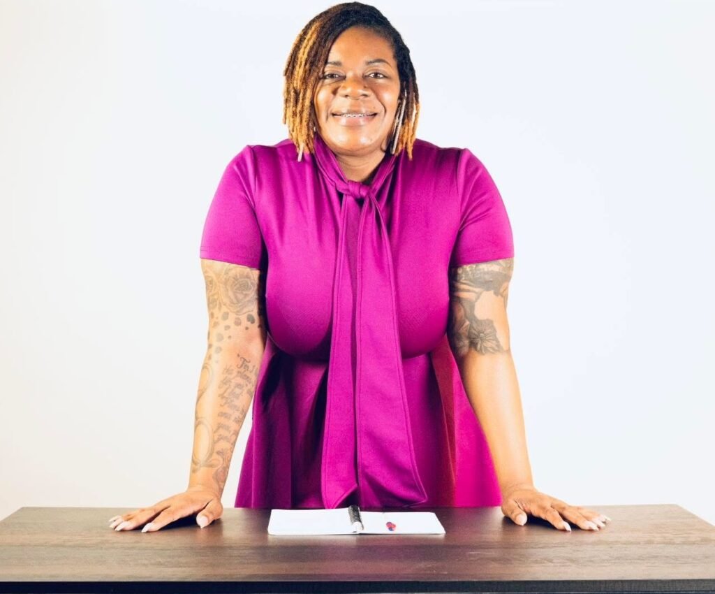 The founder, Tenisha Paige in a magenta shirt standing confidently at desk with laptop in modern office
